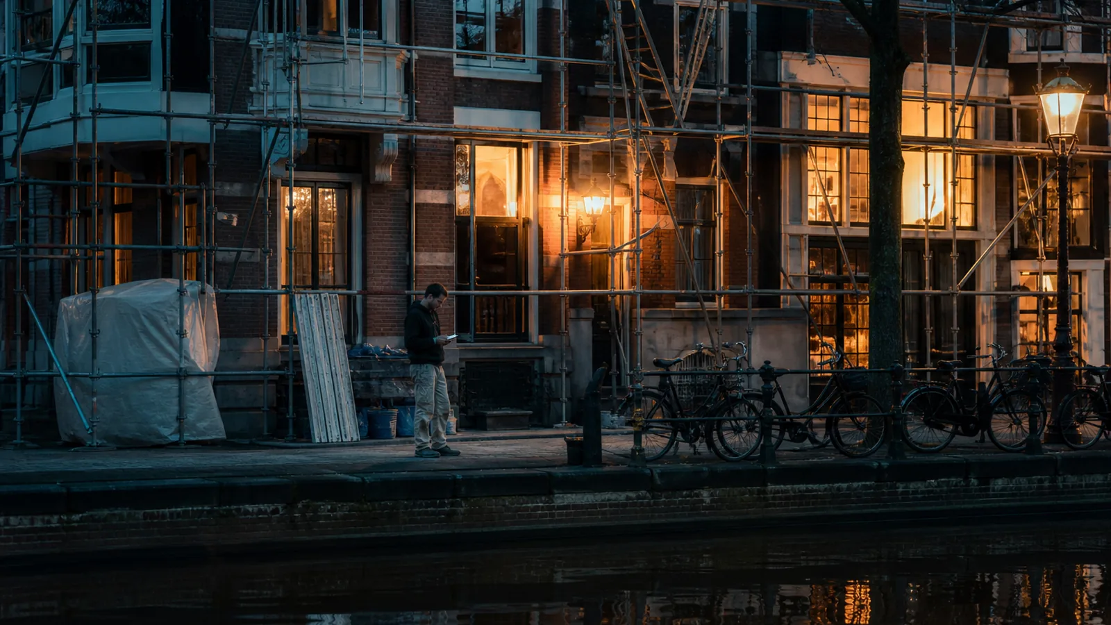 Amsterdam canal house under renovation at dusk, scaffolding and bicycles, worker checking a phone, warm window light
