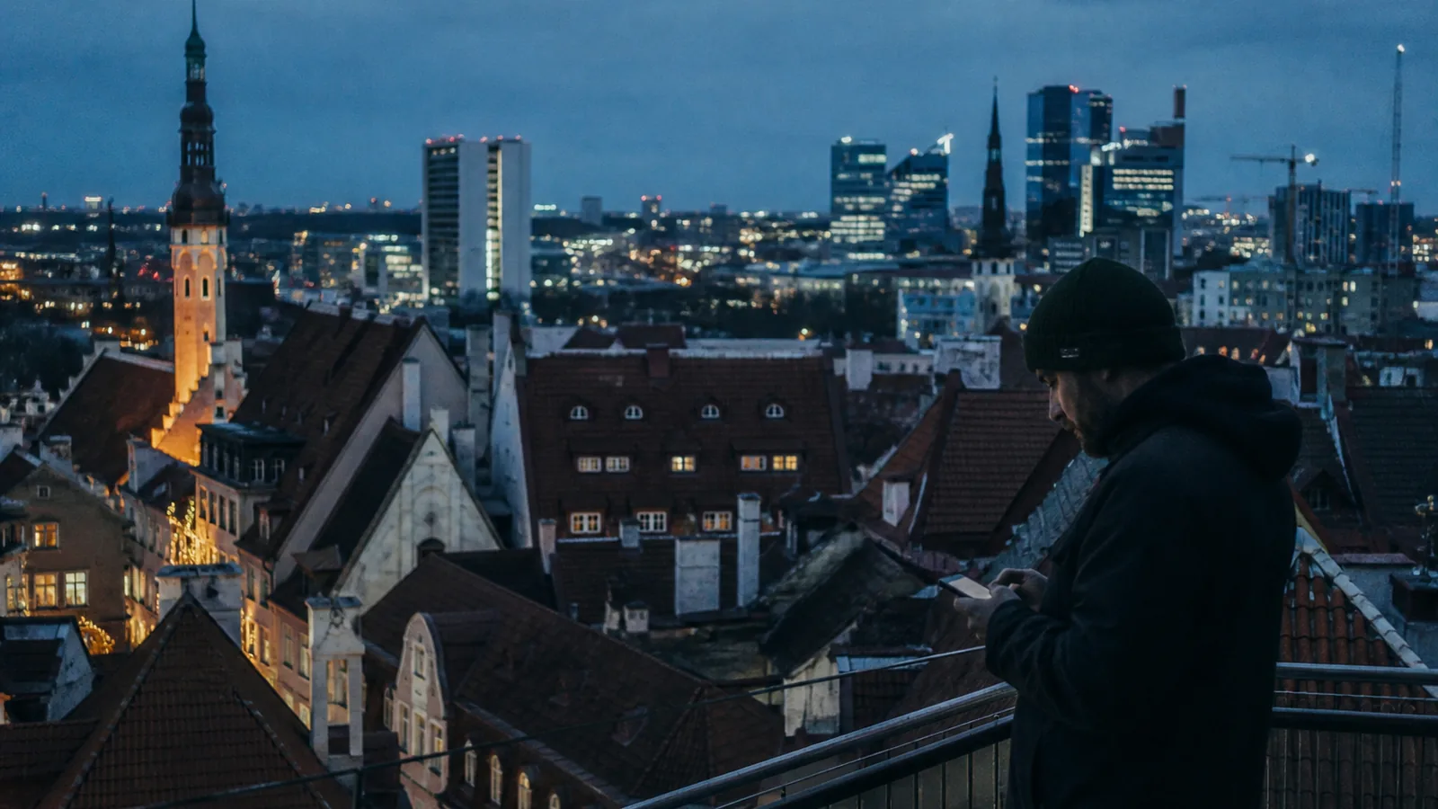 Tallinn old town at blue hour with a modern office district behind, a worker checking a phone, cool northern light with